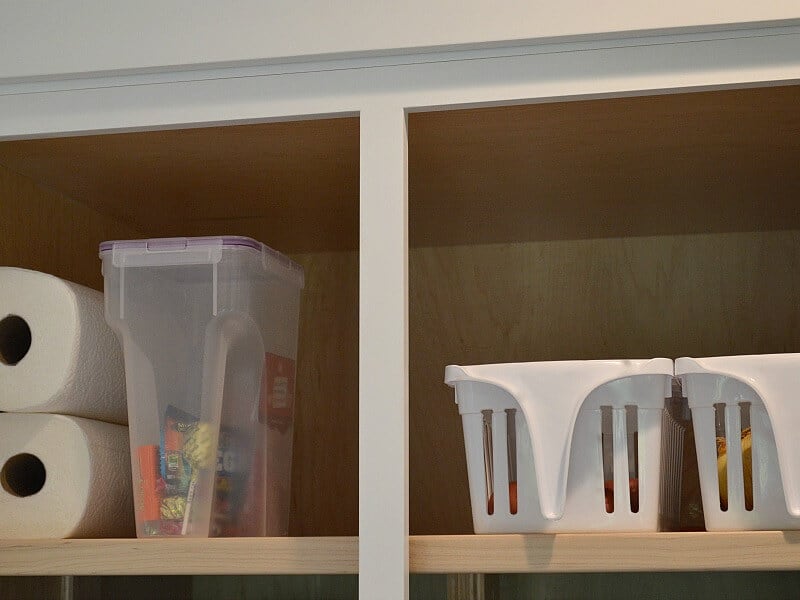 one shelf of pantry of stacked paper towel rolls, container of candy and 2 white handled baskets.