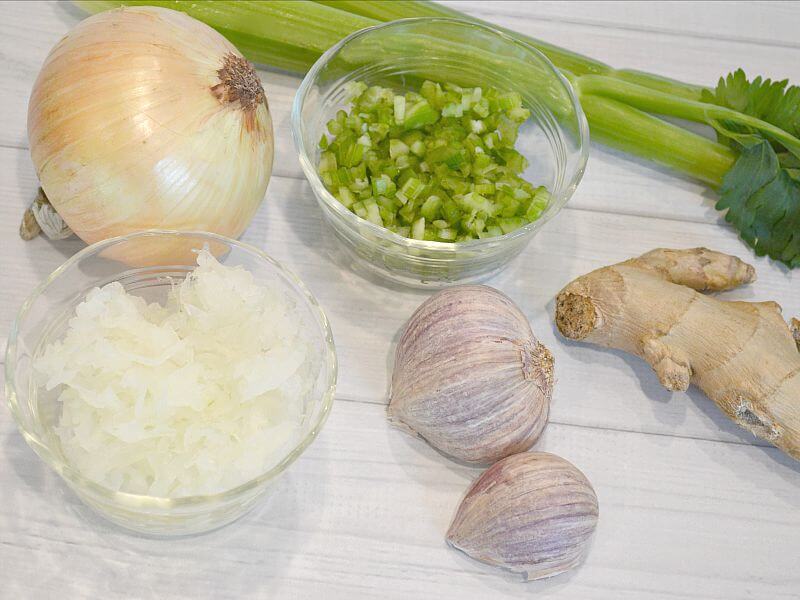 overhead view of onion, garlic, ginger root, bowl of chopped onion, bowl of chopped celery and celery stalk on white wood table