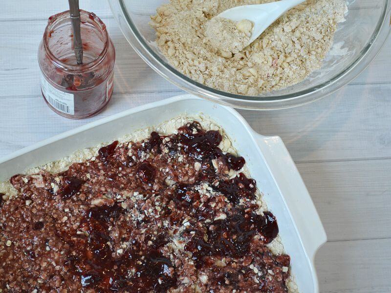 bowl of baked bars dough, jar of berry jam and baking pan of dough with layer of berry jam