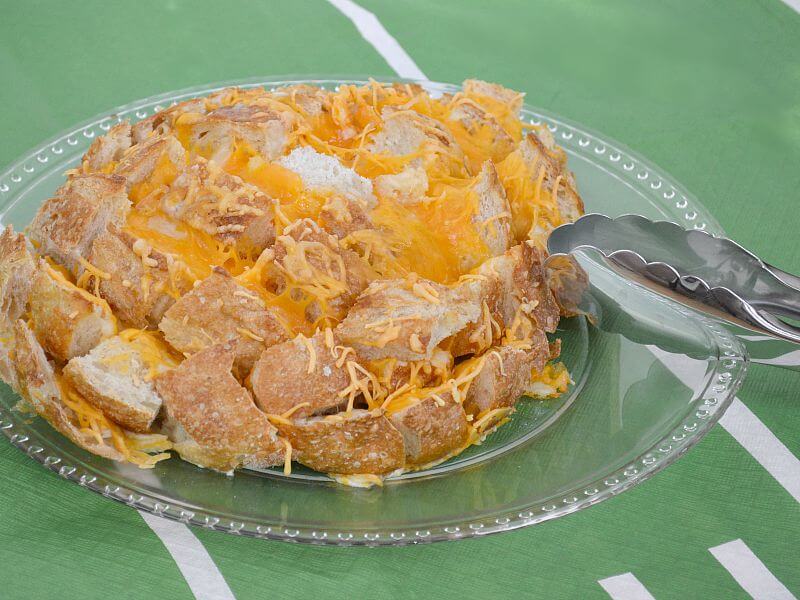 large loaf of crusty bread with melted cheese on clear plate with silver tongs on green table cloth