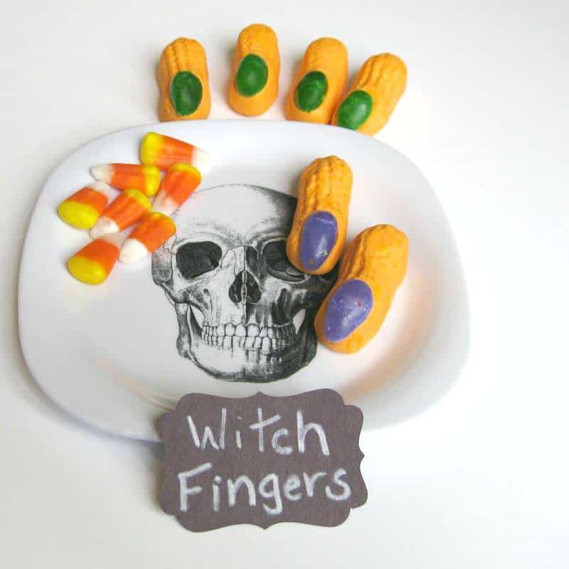white plate with black skull image holding a pile of candy corn and 2 orange circus peanuts with purple jelly bean nails with more candy fingers in the background with green candy nails and black sign with white text saying "witch fingers"