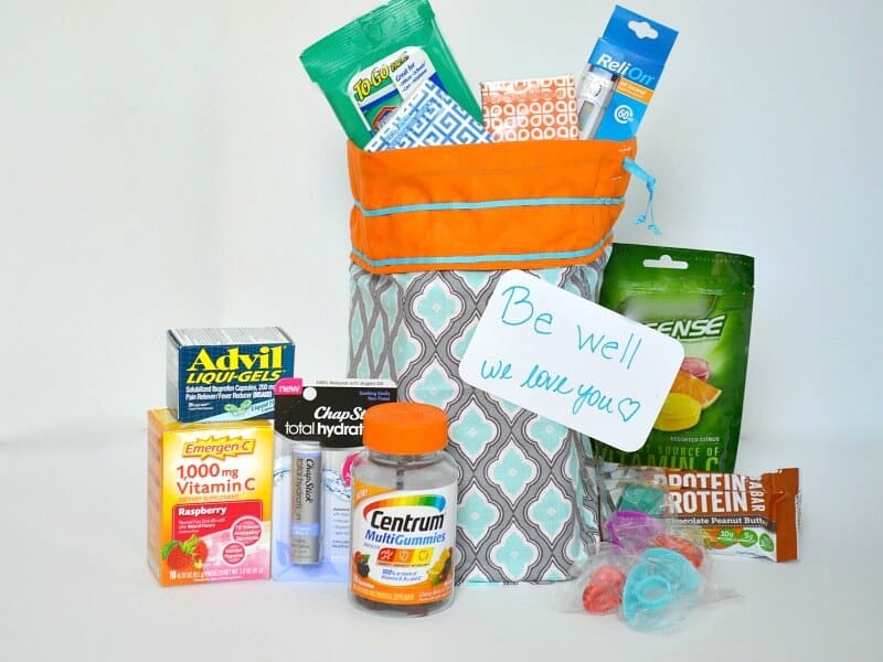 blue and orange gift bag filled with packages of tissues and a thermometer with bottle of vitamins, tube of lip balm, boxes of medicine on white table
