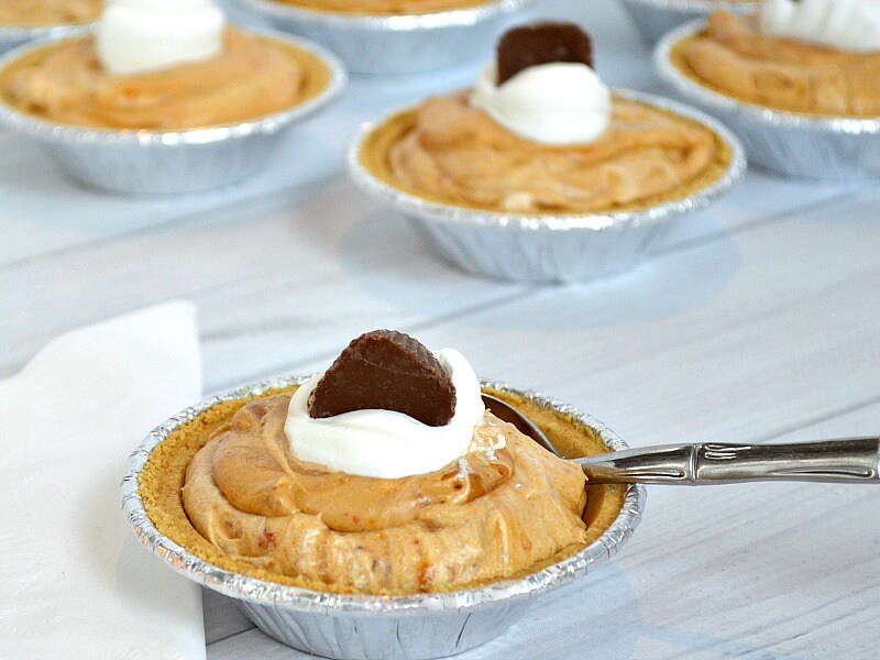 close up of small pies with light brown filling, small dollop of whipped cream topped with small piece of chocolate on white wood table and spoon in one pie crust