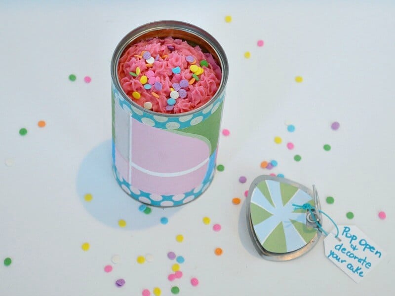 overhead view of decorated tin can opened to show frosted pink cupcake and colorful sprinkles on white table