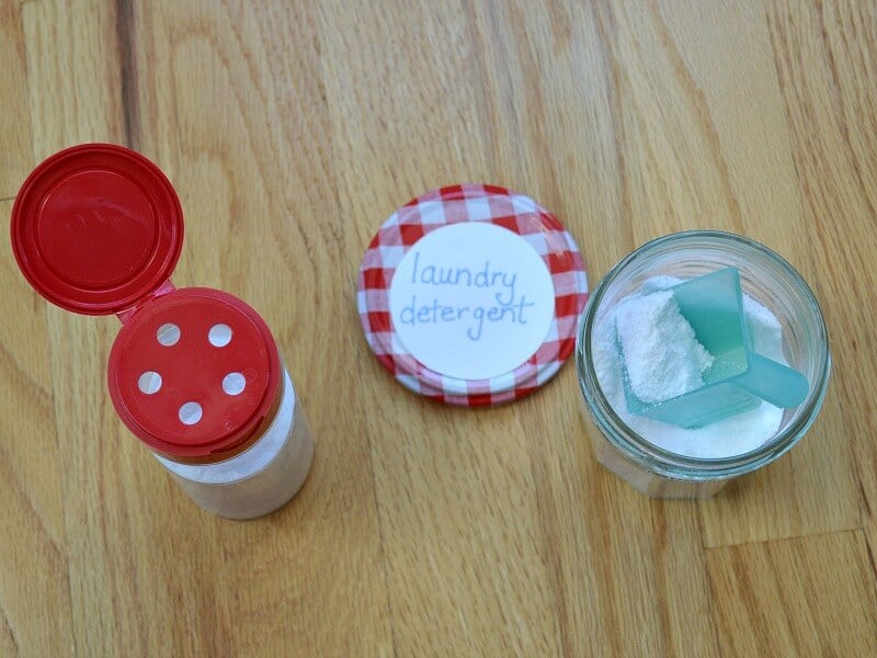 overhead view of bottle with red shaker lid next to glass jar with small blue scoop and red checked lid that says "laundry detergent"