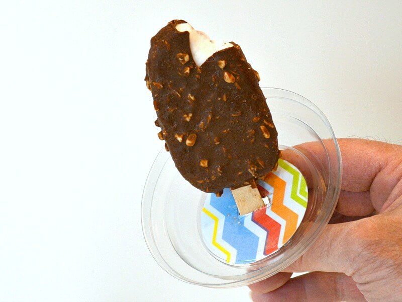 hand holding wood stick of ice cream bar with clear lid with decorated paper between hand and ice cream