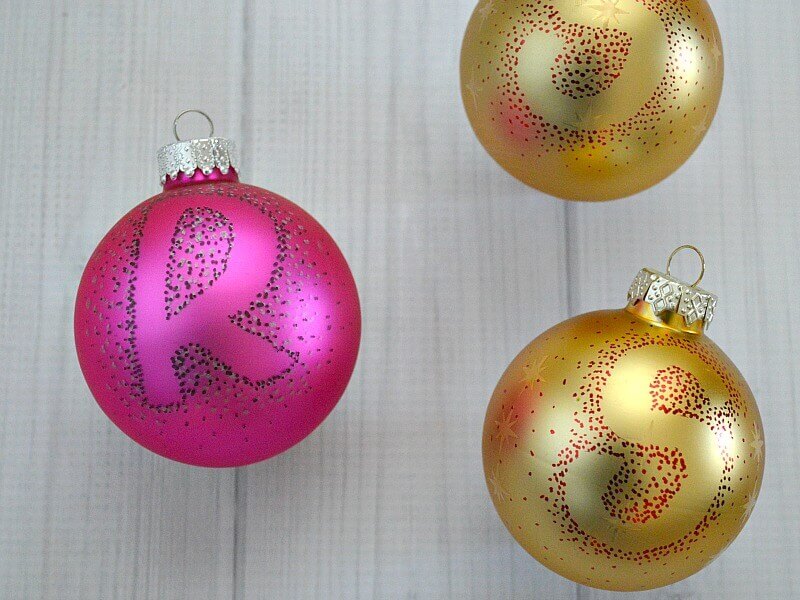 overhead view of 2 gold Christmas ball ornaments and one pink with monogrammed letters in red dots on white wood table