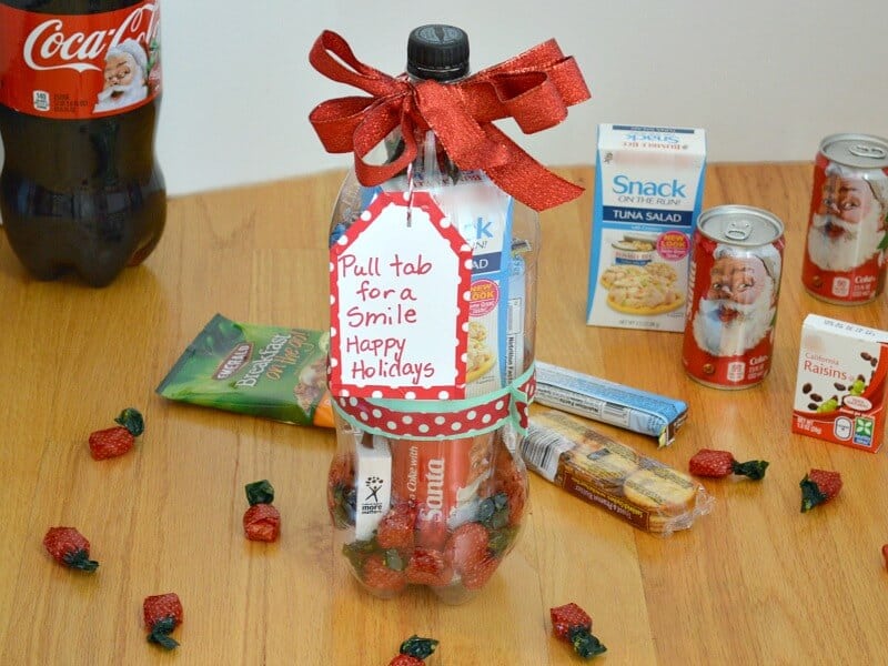 clear 2-liter soda bottle filled with treats with a red bow and tag that says "Pull tab for a smile Happy Holidays" and candy and treats on wood table