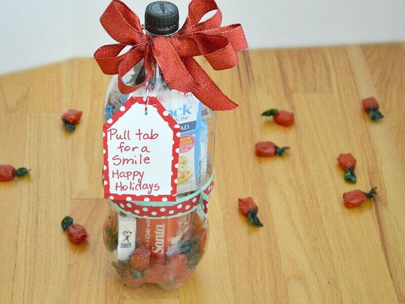 close up of clear 2-liter soda bottle filled with treats with a red bow and tag that says "Pull tab for a smile Happy Holidays" and candy scattered on wood table