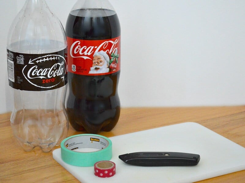 filled 2-liter bottle of Coca-cola and empty bottle next to cutting board, knife,  roll of green washi tape and roll of red polka dot washi tape