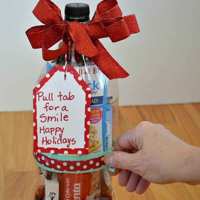 close up of clear 2-liter soda bottle filled with treats with a red bow and tag that says "Pull tab for a smile Happy Holidays" and hand holding tab