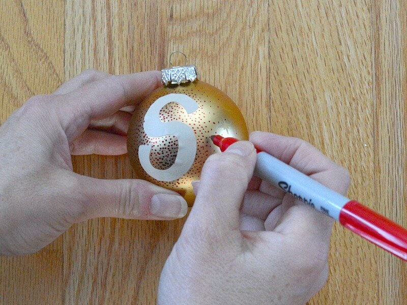 hands holding gold ball ornament and red marker making dots around a sticker S monogram on the ornament