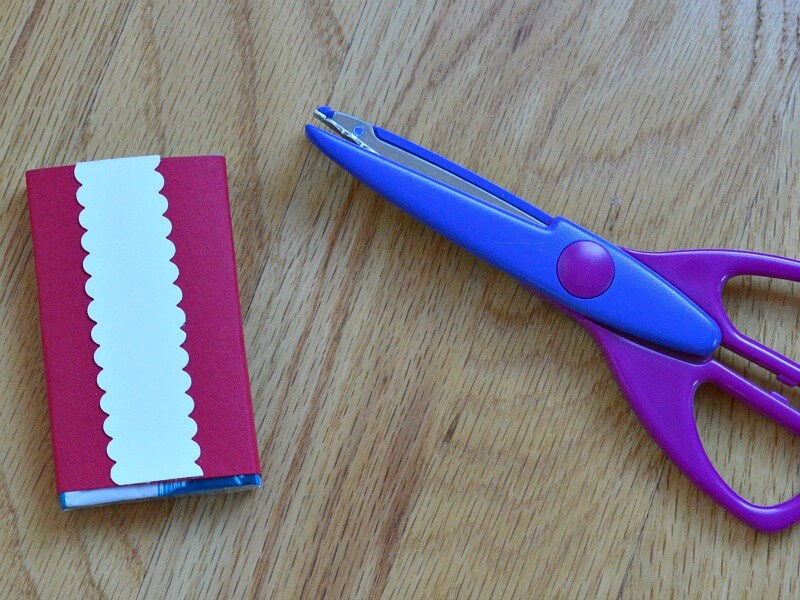 pair of purple scissors next to red box with white strip of paper with scalloped edges