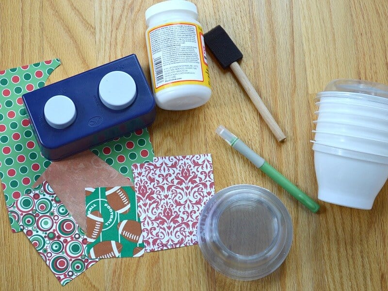 overhead view of stack of white plastic cups, stack of clear lids, marker, sponge brush, jar of glue, circle punch and various designs of decorative paper