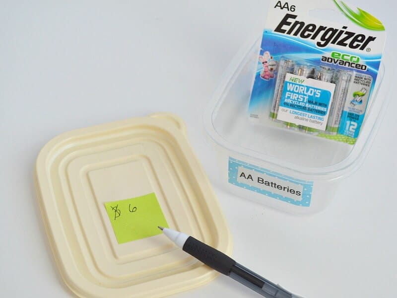 small yellow sticky note on cream lid with the number 6 written on it next to clear container with package of batteries