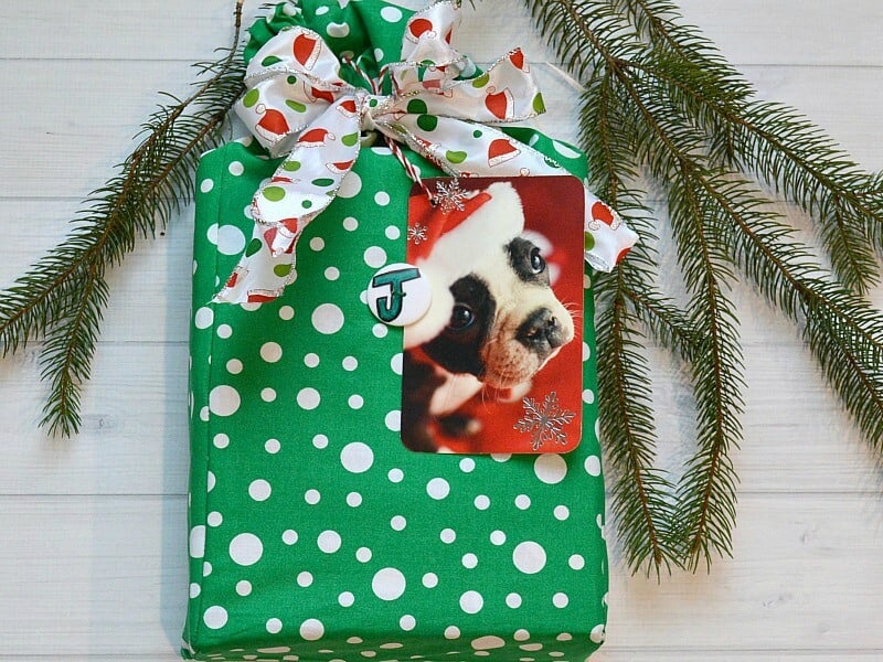 overhead view of gift with green bag and white polka dots with dog in Santa hat tag on white wood table with pine bough