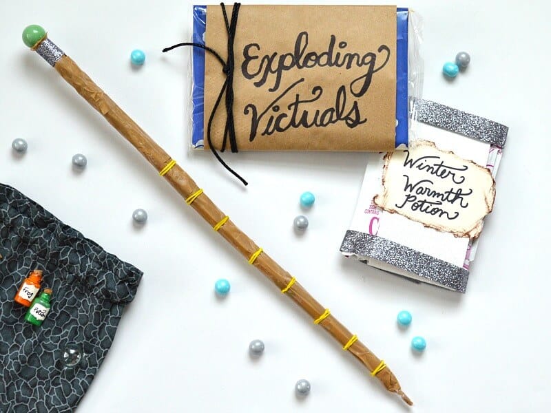 overhead view of DIY magic wand, DIY packet of "exploding victuals" and DIY packet of "Winter Warmth Potion" next to grey pouch with small glass bottles and blue and grey marbles scattered on white table