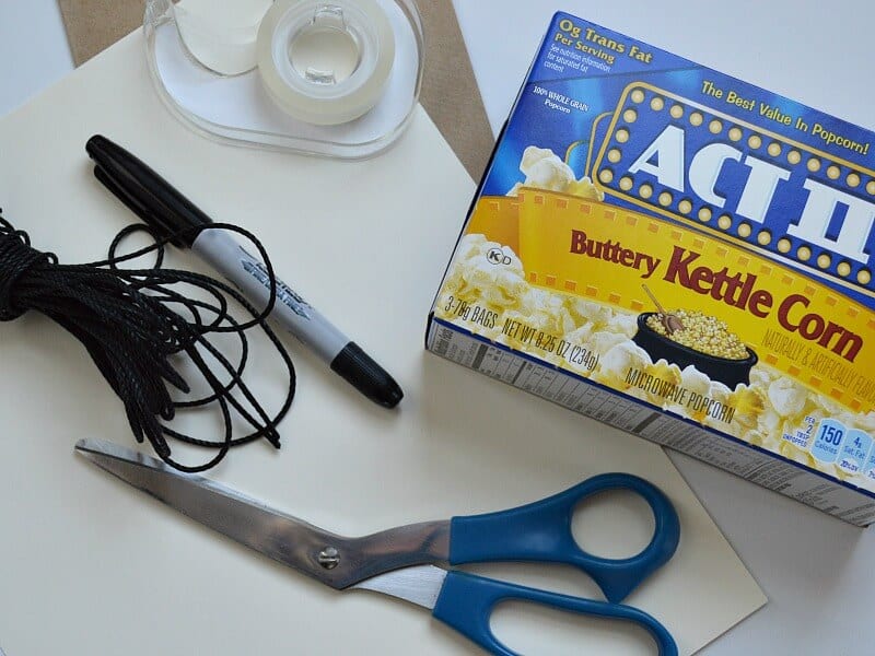 overhead view of blue and yellow box of microwave popcorn packets, blue handled scissors, black cording, roll of tape, black marker and white craft paper