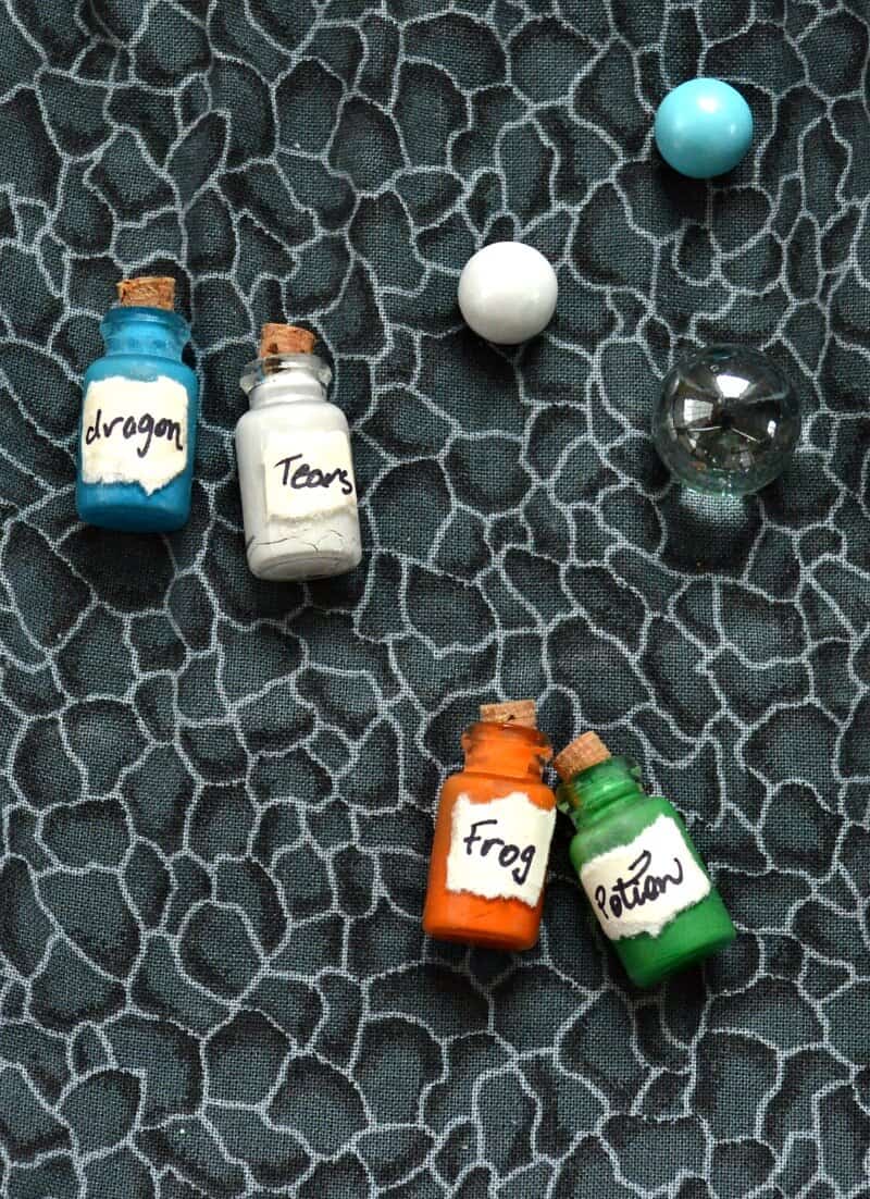 overhead view of 4 colorful tiny glass bottles with corks and handmade labels on grey fabric with blue, white and clear marble