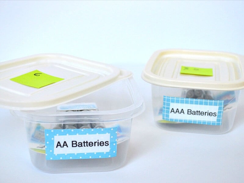 2 small clear containers with blue and white labels with names of different sizes of batteries on white table and cream lids on top of containers