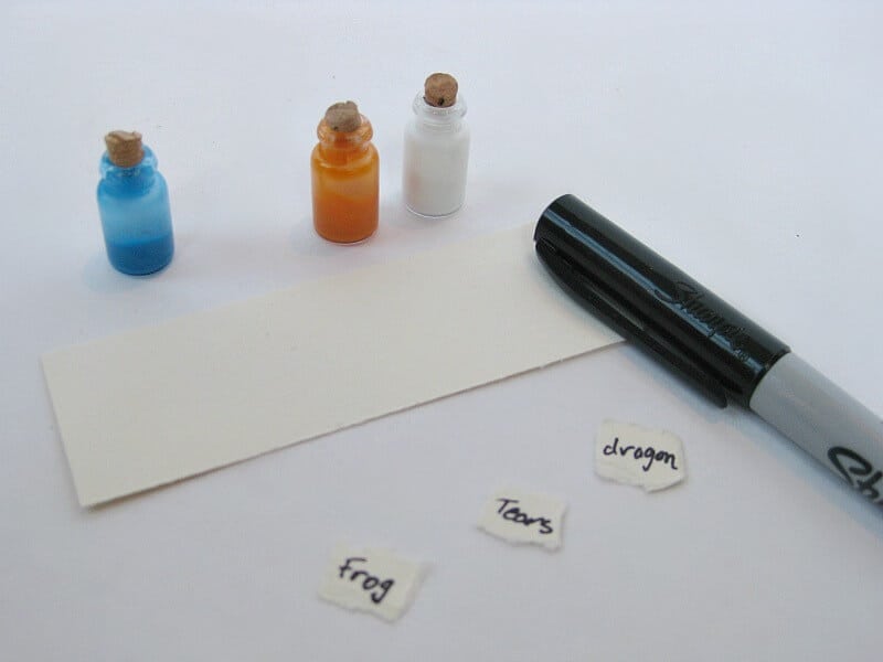 piece of cream paper, 3 small labels saying "dragon", "tears" and "frog" next to black marker and tiny blue, orange and white bottles with corks