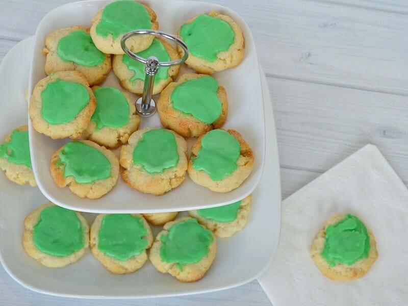 overhead view of plate of green iced cookies and one cookie on napkin on white table