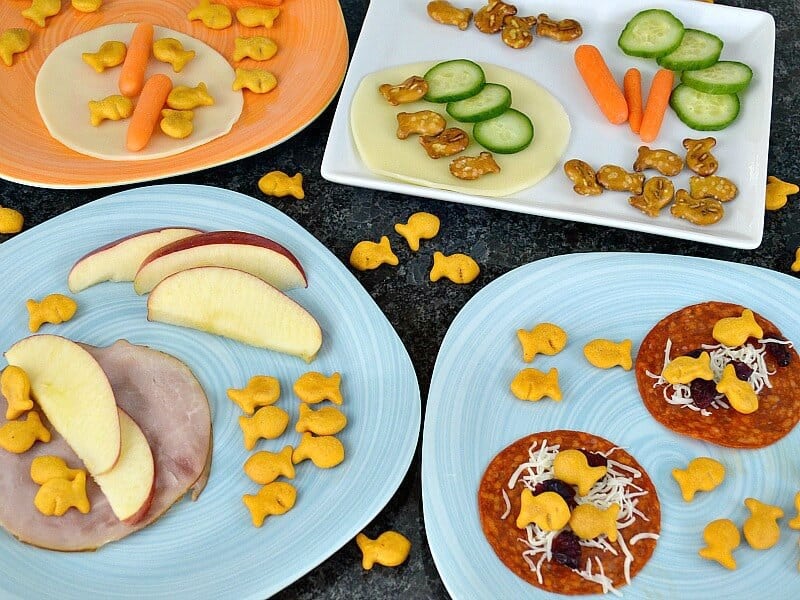 kids snacks on 4 different plates with fruit, vegetables, fish crackers and cheese stacked like open-face sandwiches