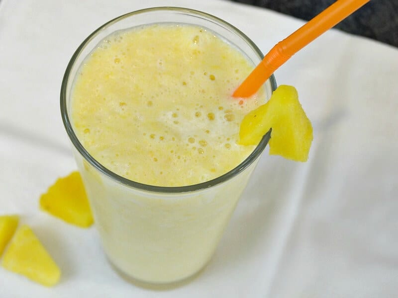 overhead view of yellow smoothie in glass with orange straw and pineapple slices on white napkin