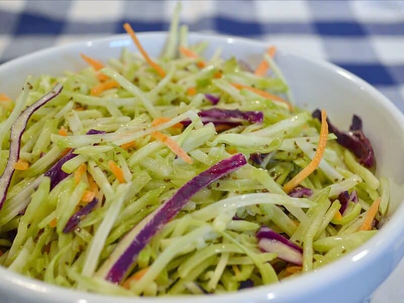 close up of green, purple and orange ingredients of coleslaw in white bowl on blue and white checked tablecloth