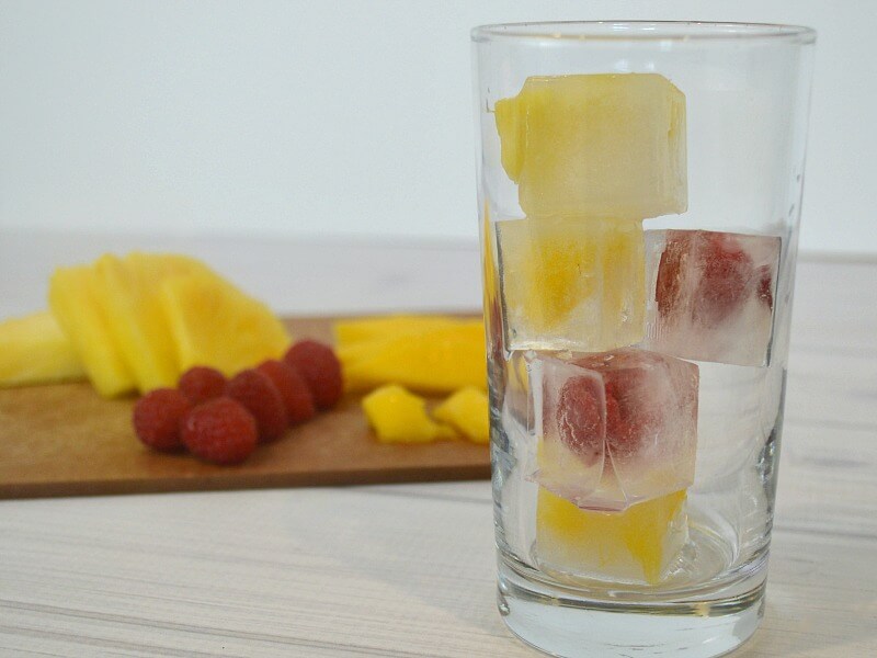 glass with ice cubes filled fruit and cutting board of fruit in background