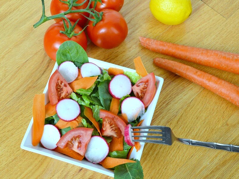 overhead view of colorful salad in white bowl with fork and vegetables on wood table