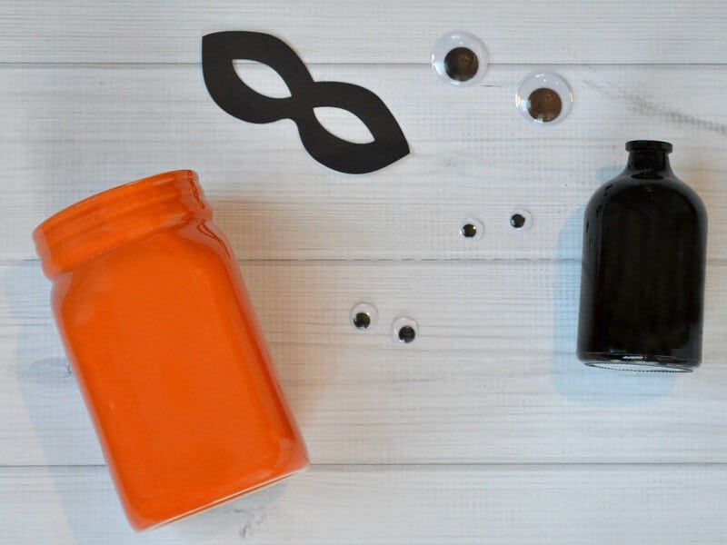 overhead view of orange jar, black jar, googly eyes and black eye mask on white wood table