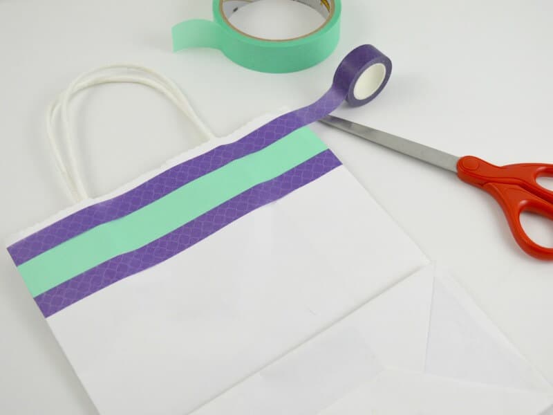 white gift bag decorated with green and purple washi tape and red scissors