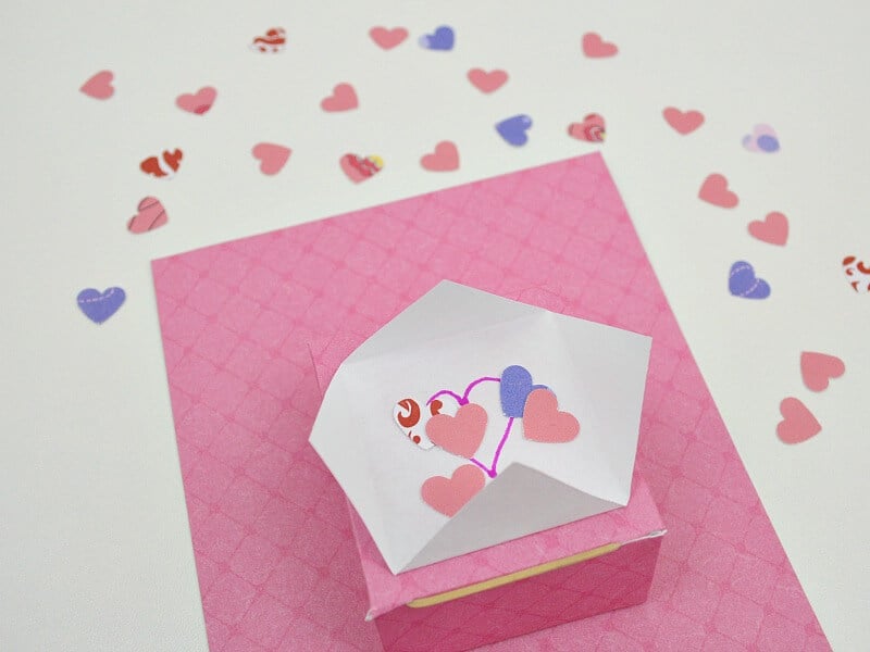 confetti flinger box on pink paper backing with pink and purple hearts scattered on table