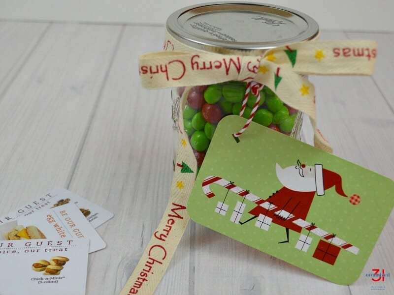 close up of clear jar of red and green candy with bow and Santa gift tag and several gift cards on table