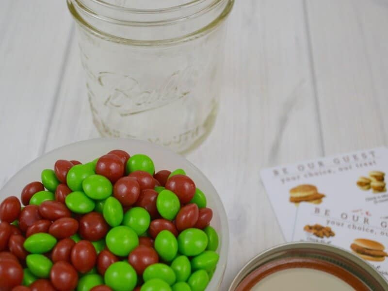 clear jar next to bowl of red and green candy and gift cards on white wood table