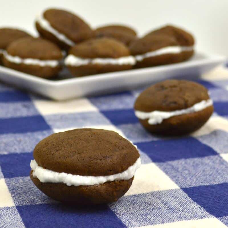 2 chocolate sandwich cookies with white filling on checked tablecloth with plate of cookies in background.