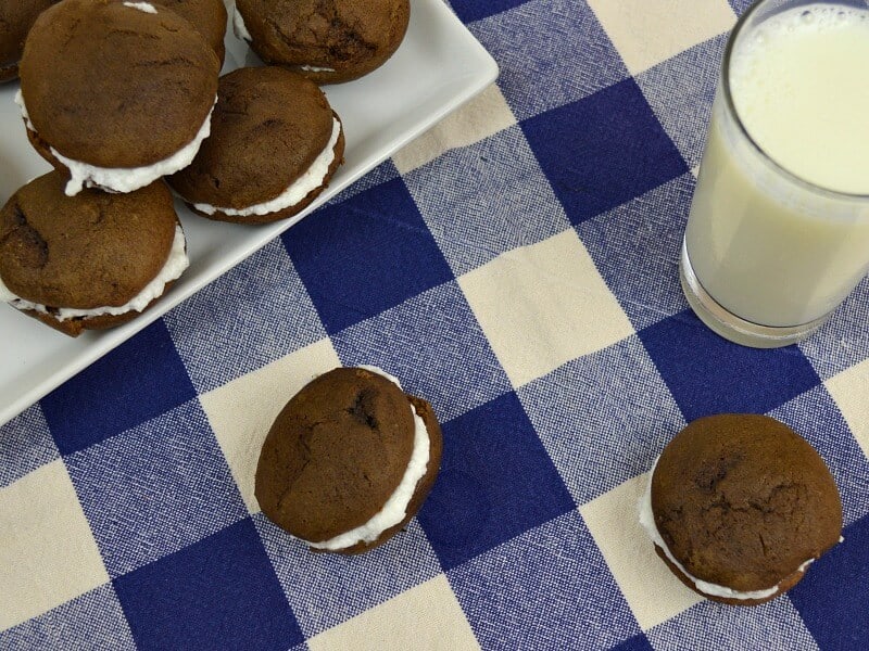 Overhead of 2 chocolate sandwich cookies on tablecloth with plate of cookies and glass of milk.