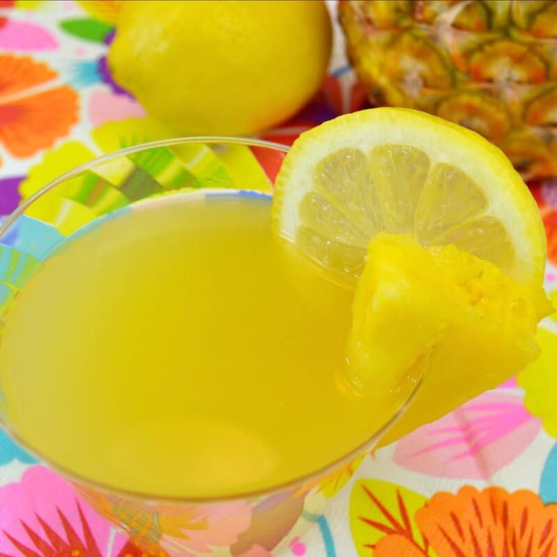 close up of yellow beverage with slice of pineapple and lemon and floral napkins on table.