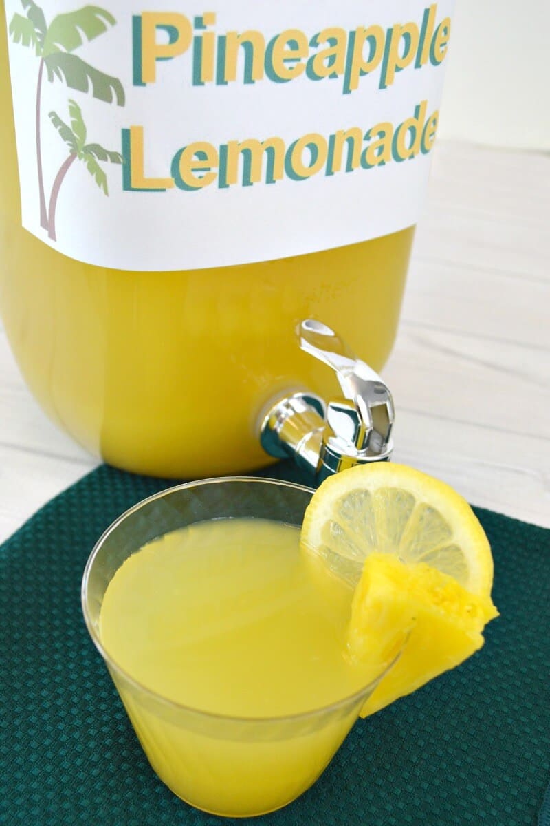 cup of yellow liquid with slice of lemon and pineapple  in front of beverage dispenser with sign "pineapple lemonade"
