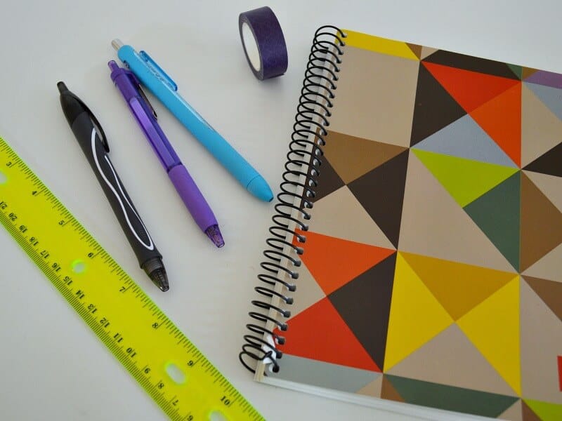 spiral notebook, yellow ruler, 3 pens and roll of washi tape on white table
