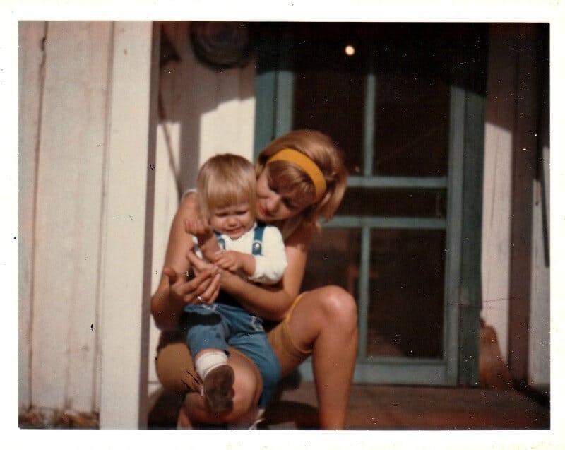 1960's image of blonde child on mom's lap looking at child's arm.