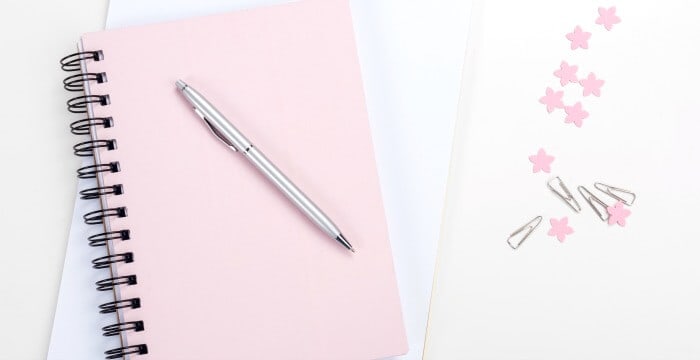 overhead view pink spiral notebook with silver pen on the cover and paperclips and pink stars on white table