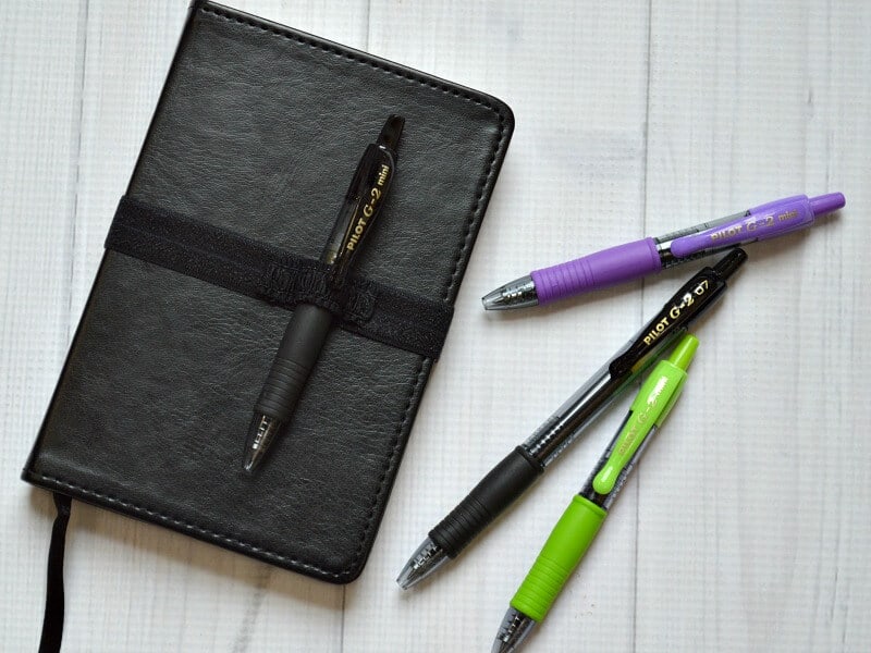 black journal with black strap holding one black pen and 3 pens on table next to it