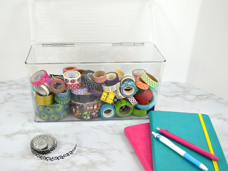 Clear bin holding colorful washi tape with 2 notebooks and pens in foreground.