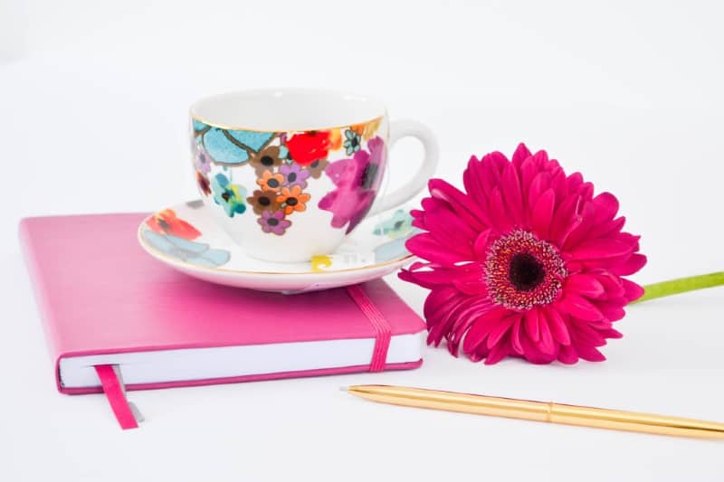 floral-pattern teacup sitting on top of pink journal next to gold pen and pink flower.