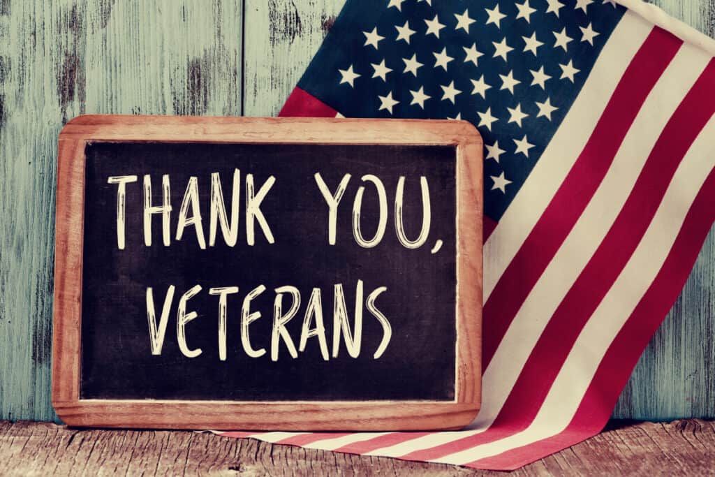 chalkboard sign with words "Thank you, veterans" on wood table in front of American flag.