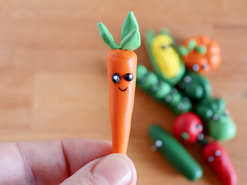 hand holding tiny clay carrot with face and other veggies in background on table