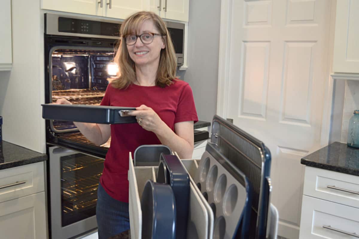 woman in red shirt holding blue baking pan next open oven and baking set in foreground