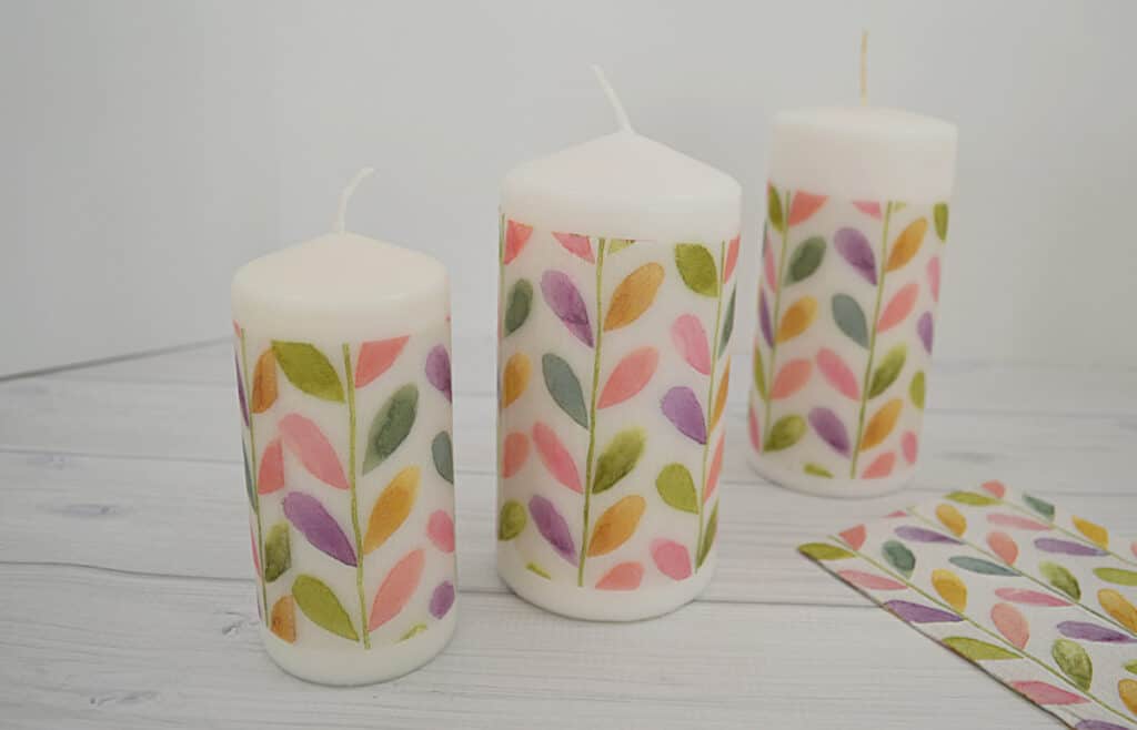 3 candles with watercolor leaves and paper towels with same design on white wood table.
